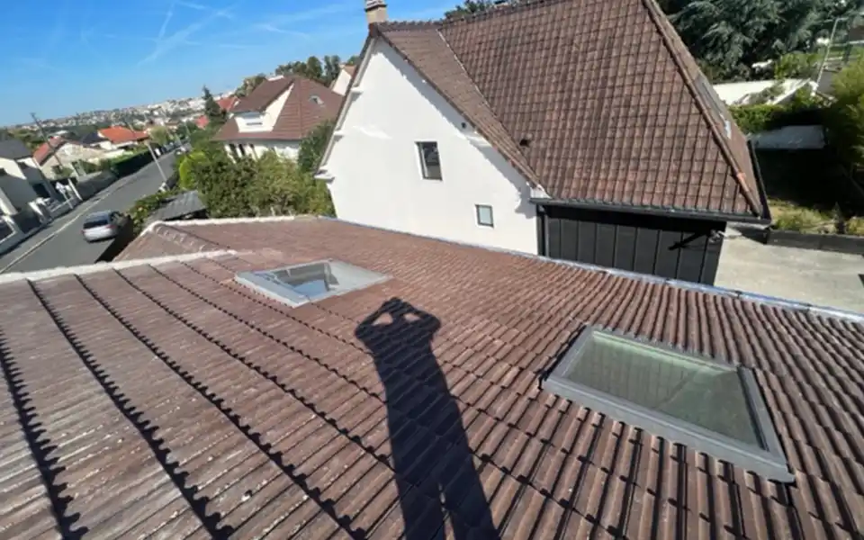 Ombre d'une personne prenant une photo sur un toit de tuiles brunes avec deux lucarnes, surplombant un quartier de banlieue - rappel de l'importance d'entretenir sa toiture dans le Loir-et-Cher pour pr&eacute;server la beaut&eacute; et la s&eacute;curit&eacute; de son habitat.