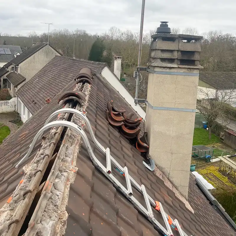 Réparation de toiture 41 Un toit de maison avec des tuiles manquantes et cassées le long du faîtage ; plusieurs tuiles et chemins de câbles sont empilés sur la surface inclinée près d'une grande cheminée, ce qui suggère une réparation de toiture en cours dans le 41.